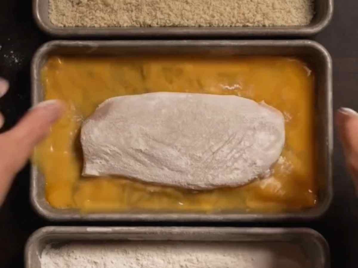 - Mão empanando os peitos de frango na farinha panko crocante para fritar.